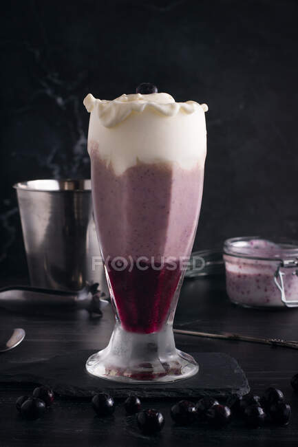 Glass of tasty smooth banana and blueberry drink with whipped cream on top on cutting board on dark background — Stock Photo