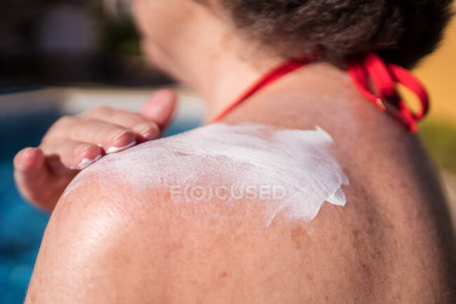 Side view of anonymous carefree female with gray hair applying sunblock on shoulder while enjoying sunny day on poolside — Stock Photo