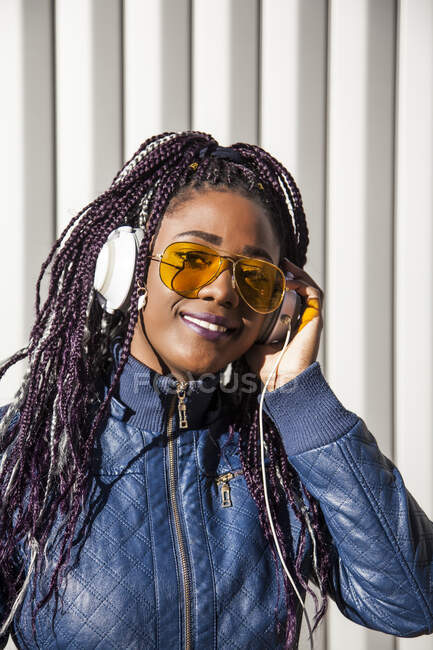 Fröhliche junge Afroamerikanerin mit Afro-Zöpfen in blauer Jacke und stylischer Sonnenbrille, die Musik über Kopfhörer genießt, während sie im Sonnenlicht vor gestreifter Wand chillt — Stockfoto
