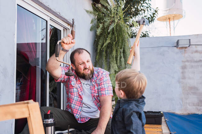 Bearded cheerful dad teaching son with hammer working with wood while sitting on boardwalk on weekend — Stock Photo