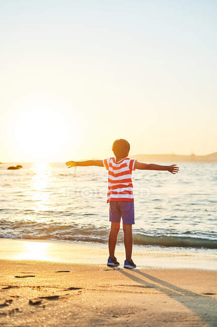 Back view full length of unrecognizable boy standing with outstretch arms on wet sandy shore washed by waving blue sea at sundown — Stock Photo