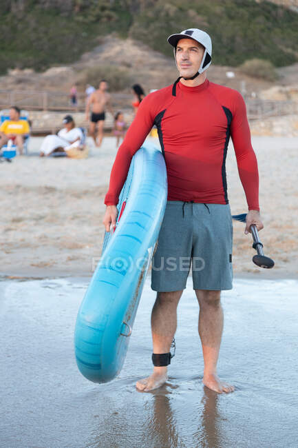 Hombre surfista en traje de neopreno y sombrero de pie mirando hacia otro lado con tabla SUP mientras se prepara para surfear en la orilla del mar - foto de stock