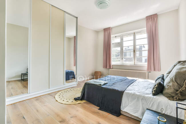 Интерьер современной светлой спальни с удобной кроватью с подушками и шкафом с зеркальной поверхностью, расположенной возле окна — стоковое фото
