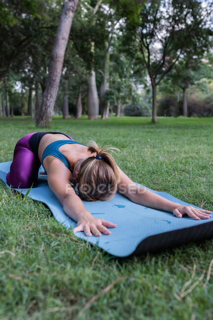 Mujer joven irreconocible en ropa deportiva que se estira mientras practica yoga en la estera sobre hierba verde en el parque a la luz del día - foto de stock