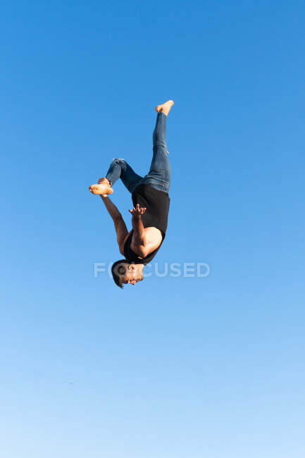 Visão lateral de baixo ângulo do atleta masculino ativo fazendo backbend enquanto salta contra palmeiras sob o céu azul sob a luz solar — Fotografia de Stock
