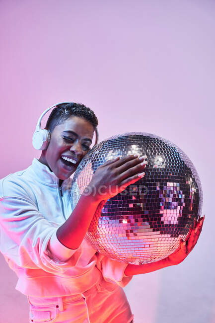 Coole ethnisch fröhliche Frau mit kurzen Haaren in drahtlosem Headset und trendiger Kleidung, die Hip-Hop mit offenem Mund und geschlossenen Augen tanzt und Glitzerball auf violettem Hintergrund hält — Stockfoto