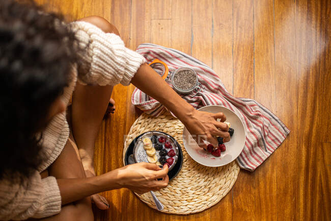 High angle of crop anonymous female sitting with crossed legs and eating sweet sliced bananas blueberry and raspberries — Stock Photo