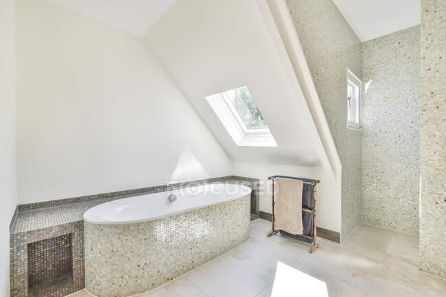 Bañera con azulejos de mosaico contra bastidor con toallas y ventanas en baño contemporáneo en día soleado - foto de stock