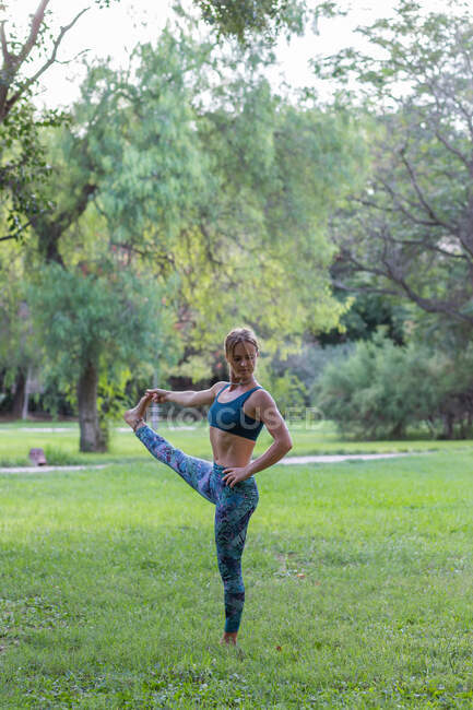 Все тело гибкой женщины в активной одежде делает Utthita Hasta Padangushthasana во время практики йоги в зеленом парке в дневное время — стоковое фото