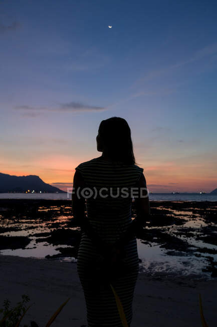 Вид сзади силуэт неузнаваемой женщины-путешественницы, стоящей на песчаном побережье, покрытом водорослями и любующейся закатом в Малайзии — стоковое фото