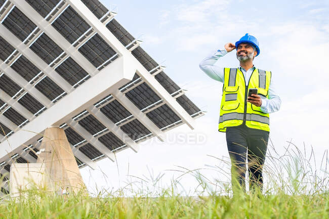 Sonriente capataz hispano de mediana edad en hardhat y chaleco navegando en el teléfono inteligente mirando hacia otro lado mientras está cerca de la estación de energía solar - foto de stock