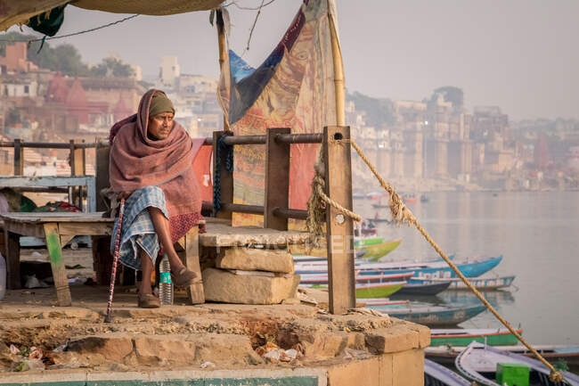 INDE, VARANASI - 27 NOVEMBRE 2015 : Homme ethnique assis sur un banc en bois près d'un bateau sur un remblai en Inde — Photo de stock
