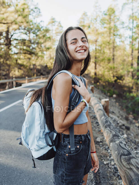 Vista lateral de mujer viajera feliz con mochila mirando a la cámara mientras camina por la carretera de asfalto en Tenerife Islas Canarias España - foto de stock