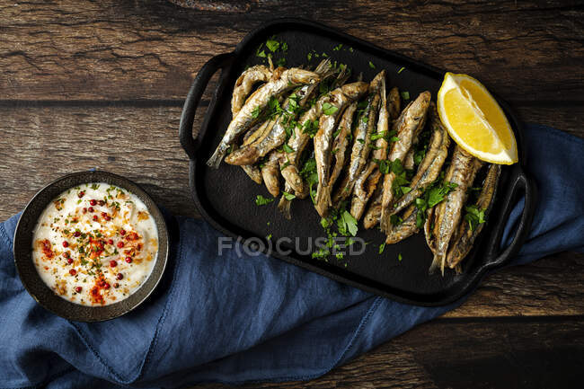 Vue du dessus de savoureux anchois frits avec morceau de citron juteux et persil haché sur plateau contre la sauce salée — Photo de stock
