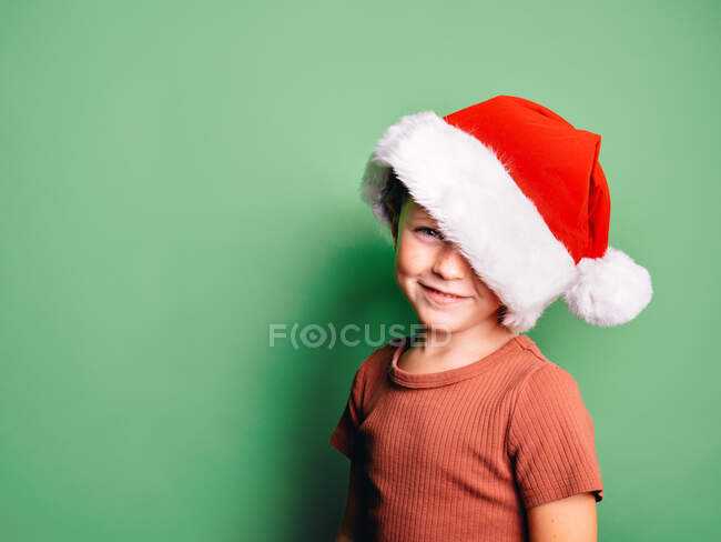 Positive small boy wearing red Santa hat smiling widely against green background and looking at camera — Stock Photo