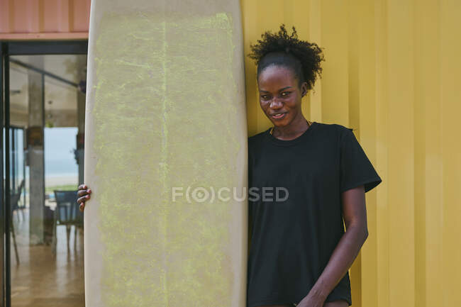 Веселая юная афроамериканская спортсменка в бикини и футболке смотрит в камеру с доской для серфинга в пляжном баре на побережье — стоковое фото