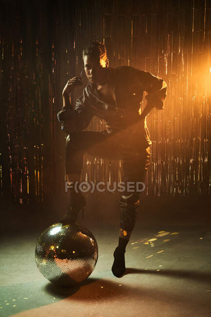 Stylish young African American woman in high heeled boots with hand on hip looking at camera while leaning on glitter ball — Stock Photo
