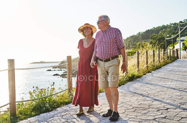 Cuerpo completo de pareja de enamorados tomados de la mano mientras pasean por el paseo marítimo pavimentado y disfrutan del mar - foto de stock