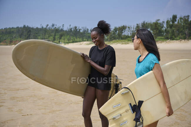 Sorrindo esportista preto com longboard contra namorada asiática com prancha de surf olhando para a frente no oceano sob céu azul nublado — Fotografia de Stock