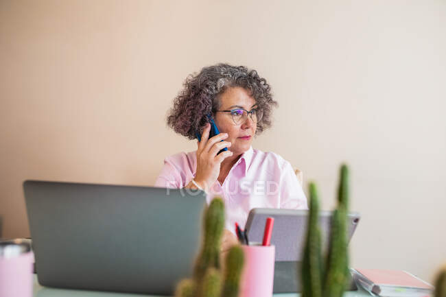 Фокусований літня жінка-підприємець в окулярах, що говорить на мобільному телефоні проти нетбука на столі в робочому просторі — стокове фото