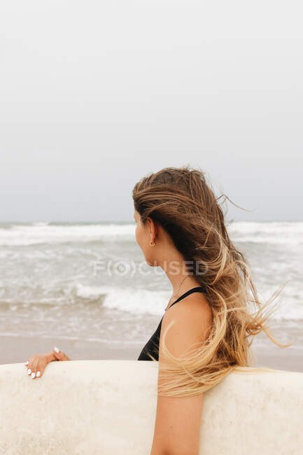 Vista lateral de una joven deportista irreconocible en traje de baño con tabla de surf mirando hacia la costa arenosa contra el océano tormentoso - foto de stock
