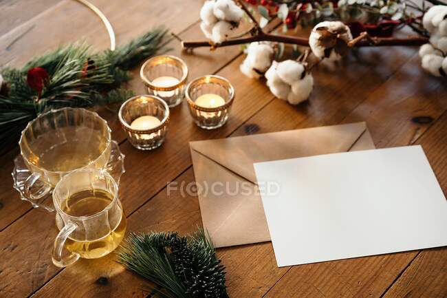 Сверху рождественской композиции с горящими свечами и чашками чая, помещенными рядом с пустой открыткой на деревянном столе, украшенном еловыми ветвями и хлопковой веточкой — стоковое фото
