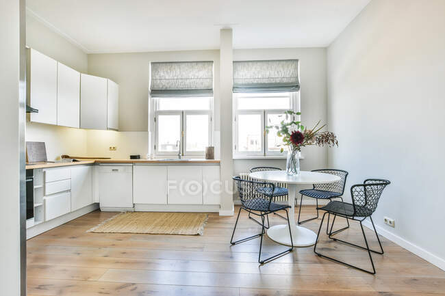 Amplia cocina luminosa con armarios blancos y zona de comedor amueblada con mesa blanca y sillas cerca de la ventana - foto de stock