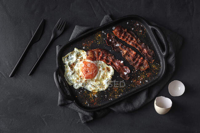 Vista superior do lado ensolarado ovo com fatias de bacon frito e condimentos na bandeja contra talheres no fundo escuro — Fotografia de Stock