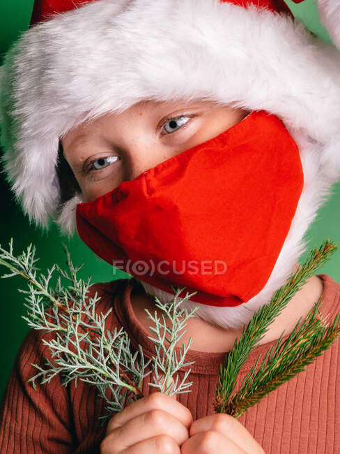 Boy in red Santa hat and mask for protecting from COVID with fir branches looking down with eyes wide open — Stock Photo