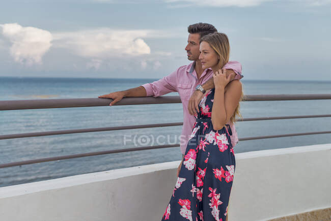 Unshaven man in wristwatch embracing contemplative female beloved in sundress on embankment against sea during travel — Stock Photo