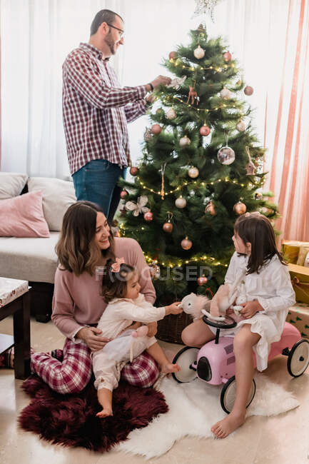 Lächelnde Mutter sitzt mit Töchtern auf Teppich und spielt mit Spielzeug neben Vater, der Weihnachtsbaum schmückt — Stockfoto