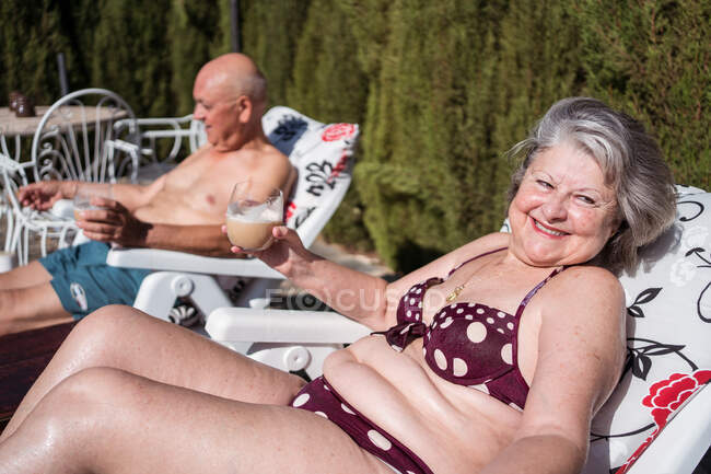 Vieux couple pieds nus en maillot de bain boire du café glacé tout en refroidissant sur des chaises longues dans la cour arrière — Photo de stock