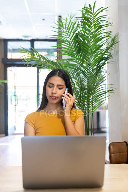 Freelancer feminino sério sentado à mesa de madeira no café e ter telefonema enquanto digita no netbook — Fotografia de Stock