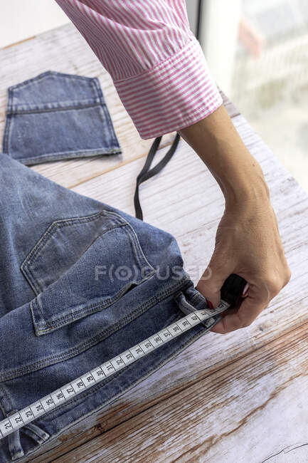 High angle of crop anonymous female dressmaker measuring waist of jeans on wooden table in daytime — Stock Photo