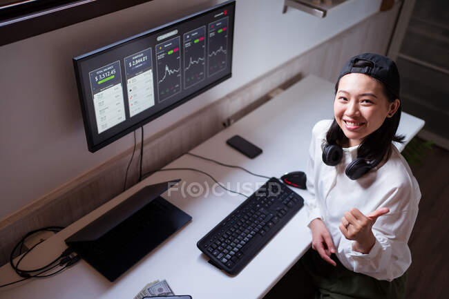 Dall'alto sorridente broker etnico femminile digitando sulla tastiera contro monitor con grafica e mostrando come gesto mentre si guarda la fotocamera a casa — Foto stock
