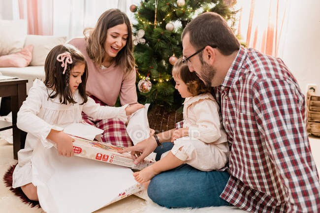 Garotas surpresas com pais alegres abrindo caixa de presente no chão enquanto celebra o dia de Natal na sala da casa — Fotografia de Stock
