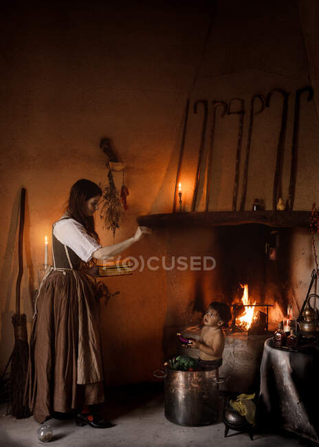 Seitenansicht der Hexe mit magischen Buch Zauber auf Kind sitzen in Metalltopf in düsteren Raum mit brennendem Kamin — Stockfoto