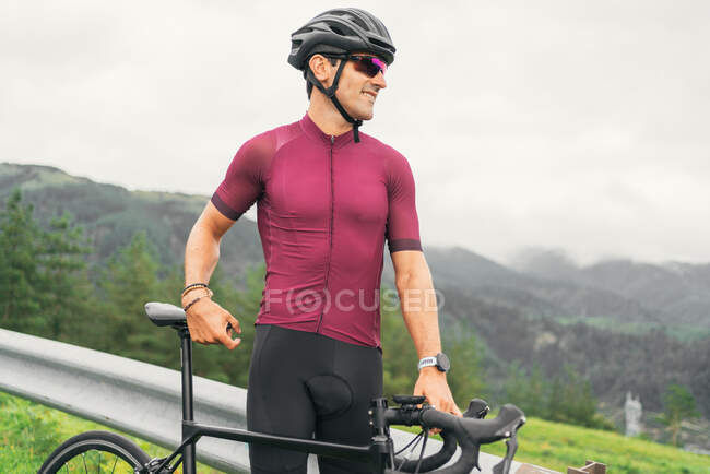 Vista lateral do desportista adulto alegre em óculos de sol de ciclismo e capacete sentado na bicicleta de estrada na estrada rural à luz do dia — Fotografia de Stock