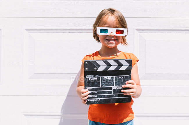 Jolie fille heureuse dans des vêtements colorés décontractés tenant clapperboard tandis que sur des lunettes tridimensionnelles debout sur fond de mur blanc — Photo de stock