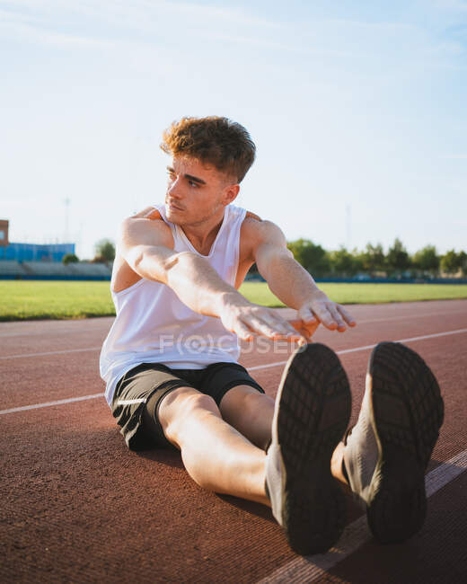 Fit male athlete in sportswear leaning forward during workout on track under cloudy sky in sunlight — Stock Photo
