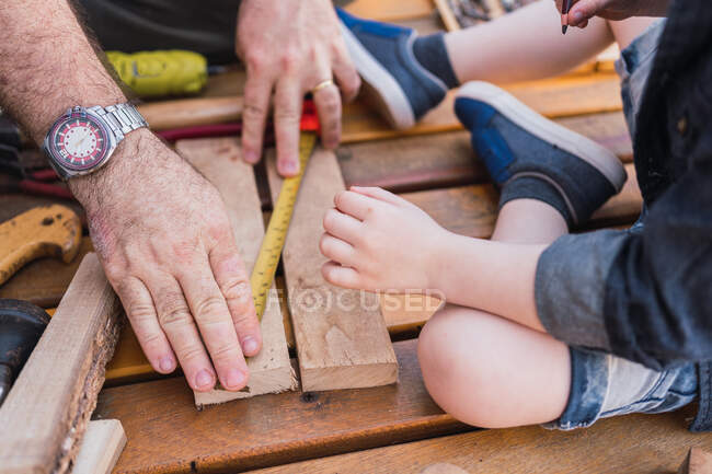 Cortar pai irreconhecível medir peça de madeira com fita contra criança com lápis sentado no calçadão — Fotografia de Stock