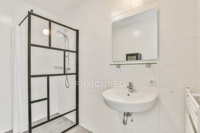Минималистский дизайн белой ванной комнаты с раковиной под зеркалом, висящей на черепичной стене рядом со стеклянной душевой кабиной в квартире — стоковое фото