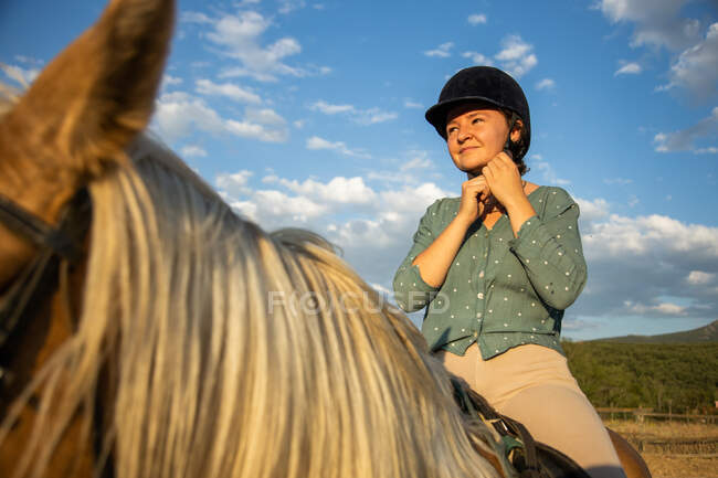 Mulher sonhadora no capacete protetor sentado na égua enquanto olha para longe sob o céu azul nublado na luz solar — Fotografia de Stock
