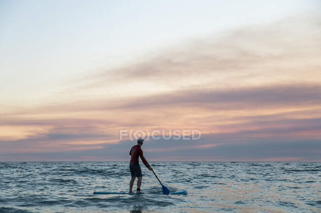 Vue arrière d'un surfeur masculin méconnaissable en combinaison et chapeau sur planche à pagaie surfant sur le rivage pendant le coucher du soleil — Photo de stock