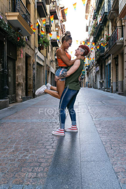 Вид збоку на модну молоду татуйовану жінку, яка носить гомосексуальну дівчину, сміючись і дивлячись один на одного на міській прогулянці — стокове фото