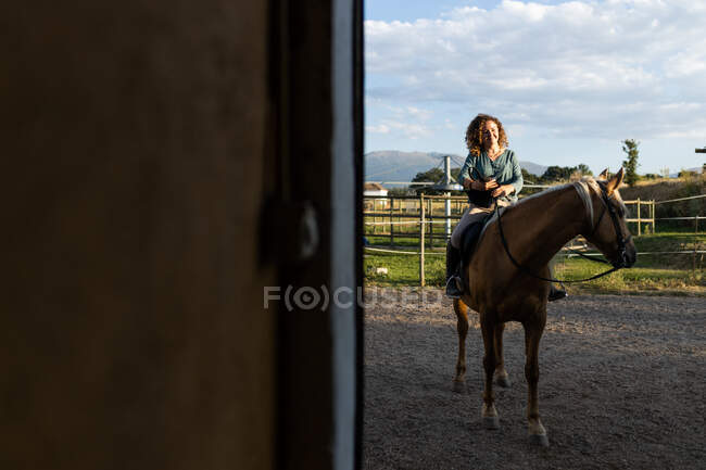 Sorrindo fêmea sentada no cavalo na tenda na escola de equitação rural — Fotografia de Stock