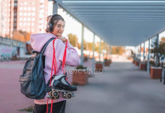 Adolescente joyeuse portant un sweat à capuche rose clair et un sac à dos avec écouteurs et patins à roulettes avec lacets roses — Photo de stock