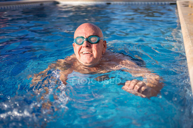 Hombre mayor relajado en gafas nadando en agua limpia transparente de la piscina mientras se relaja en el caluroso día de verano - foto de stock
