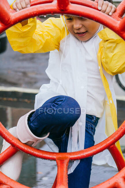 Cheerful ethnic child with wet hair in slicker having fun between metal construction in rainy weather — Stock Photo