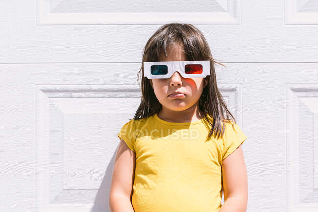 Menina bonito em roupas coloridas casuais e óculos tridimensionais em pé no fundo da parede branca olhando para a câmera — Fotografia de Stock